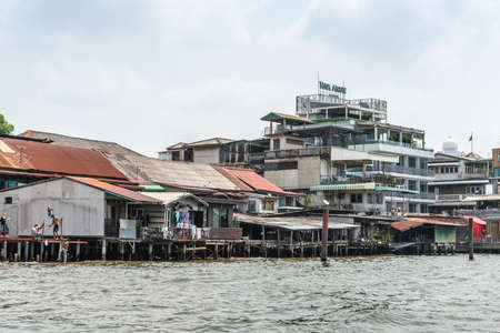 Bangkok city, Thailand - March 17, 2019: Chao Phraya River. riva Modern up to date architectural Riva Arun hotel among poor dilapidated housing on stilts along the water. People and laundry on platforms.のeditorial素材