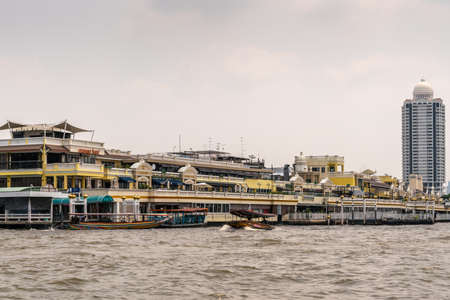 Bangkok city, Thailand - March 17, 2019: Chao Phraya River. Pak Khlong Flower Market is very wide not tall yellowish building with its own landings and plenty of other businesses under silver sky.のeditorial素材