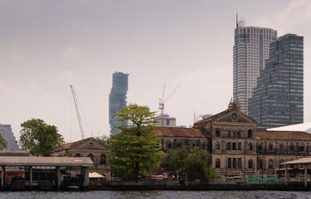 Bangkok city, Thailand - March 17, 2019: Chao Phraya River. Skyscrapers of offices and hotels around ruin of Old Customs House at quay. Silver sky with cloudscape. King Power MahaNakho skyscraper in back.のeditorial素材