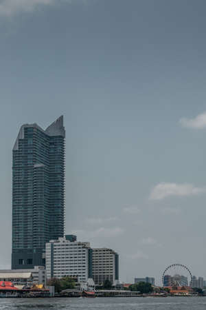 Bangkok city, Thailand - March 17, 2019: Chao Phraya River. White Ramada Plaza hotel and shopping mall with tall new Skyscraper nearby and Chang Ferris Wheel and park on side.のeditorial素材
