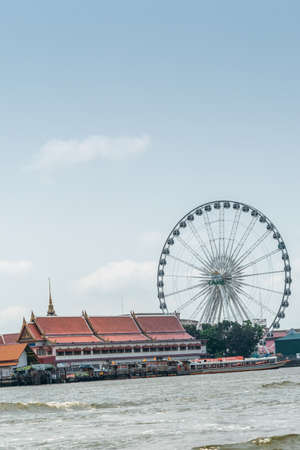 Bangkok city, Thailand - March 17, 2019: Chao Phraya River. Chang Ferris Wheel and red roofs of Wat Rajsingkorn Buddhist temple and museum in front under light blue sky. Ferry at landing.のeditorial素材