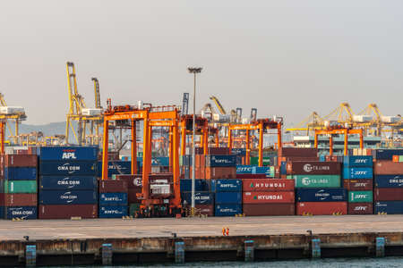 Laem Chabang seaport, Thailand - March 17, 2019: Stacks of shipping containers on quay with colorful cranes and tall orange carriers in back under silver sunset sky. Two langshoremen in front.のeditorial素材