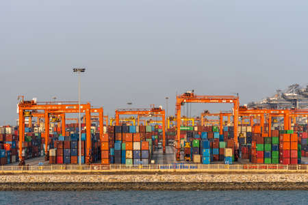 Laem Chabang seaport, Thailand - March 17, 2019: Looking through Stacks of shipping containers on quay with tall orange carriers like bridges under silver sunset sky.のeditorial素材