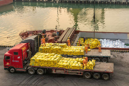 Da Nang, Vietnam - March 10, 2019: Tien Sa Port in Da Nang Bay. Men in orange garb stack manually loose yellow bags, out of ship, on 2 flat bed trucks.のeditorial素材