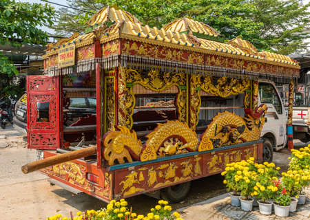 Da Nang, Vietnam - March 10, 2019: Van rebuilt as gold-red hearse with elaborate decorations of dragons and Chinese mythological symbols, among yellow flowers and green foliage.のeditorial素材