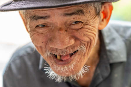 Da Nang, Vietnam - March 10, 2019: Street scene. Facial closeup of very old man who has trouble walking dressed in gray and with hat and on sandals. He is a lottery ticket seller.のeditorial素材