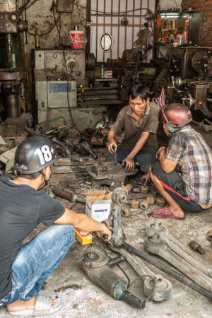 Da Nang, Vietnam - March 10, 2019: Three men, all  work in chaotic dark gray motorbike repair shop. Two of them wear helmets. Lots of machinery.のeditorial素材