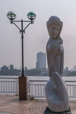 Da Nang, Vietnam - March 10, 2019: Han River Boardwalk art exhibition. Gray statue of Woman under silver sky. High buildings over river in back.のeditorial素材