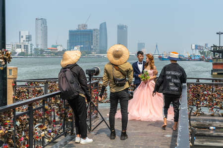 Da Nang, Vietnam - March 10, 2019: Bridal photo shoot on Love Bridge at Han River with downtown skyscrapers and fishing vessels in back. Photography crew, groom and bride in peach gown.のeditorial素材