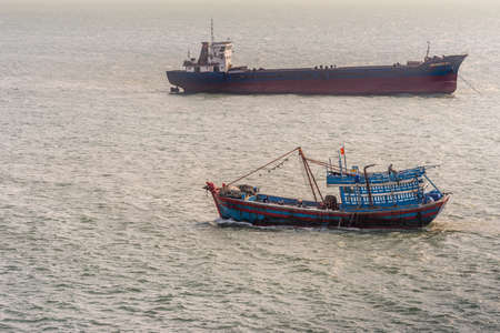 Da Nang, Vietnam - March 10, 2019: Tien Sa Port in Da Nang Bay. Black and red Binh Nguyen bulk cargo ship crosses blue and red small fishing vessel on gray bay water.のeditorial素材