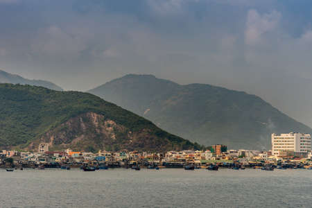 Nha Trang, Vietnam - March 11, 2019: Just north of the port, a fishing community lives pretty. much on the beach in small houses and their boats on the water. Back is tall forested mountain range.の写真素材