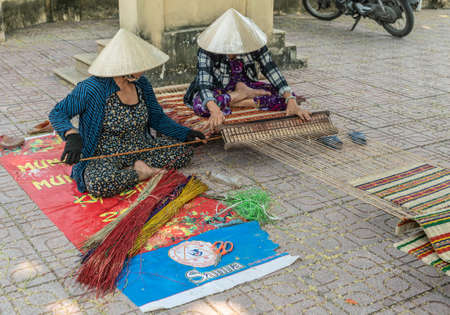 Nha Trang, Vietnam - March 11, 2019: Dinh Phu Vinh community center and celebration hall. On Courtyard, closeup of 2 women in teamwork flat weaving carpet with colored twigs.のeditorial素材