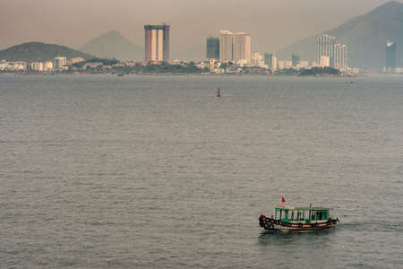 Nha Trang, Vietnam - March 11, 2019: Small brown and green ferry boat sails into harbor with skyscrapers of town in back. Foggy morning sky with tall green hills on horizon.のeditorial素材
