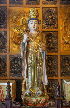 Da Nang, Vietnam - March 10, 2019: Chua An Long Chinese Buddhist Temple. Tall colorfully painted porcelain statue of royal bodhisattva with golden crown and sceptre in front of brass wall of Buddha images.のeditorial素材