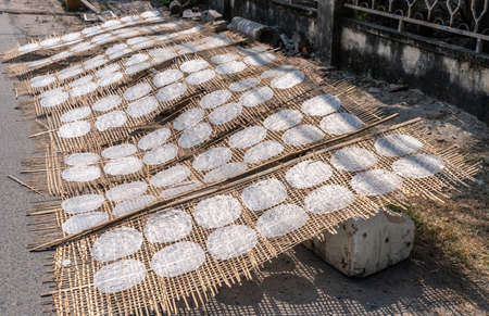 Nha Trang, Vietnam - March 11, 2019: Dai Diem Nam neighborhood. 8 large beige reed woven sheets holding each 14 white circular rice cakes exposed to sun in rural street.の写真素材