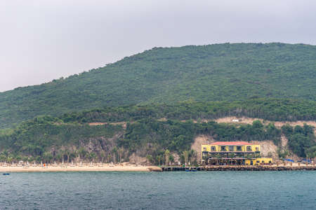 Nha Trang, Vietnam - March 11, 2019: Evening, yellow with red roof beach house with docks on Vinh Nguyen island with forested hill in back. Yellow beach, rocky cliffs and greenish water.のeditorial素材