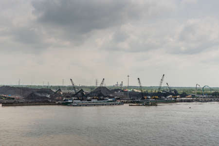 Long Tau River, Vietnam - March 12, 2019: Phuoc Khanh area. Black hills. cranes and boat docked along quay at Long Thuan Coal Port. Fine green belt of trees under dark cloudscape and brown water in front.のeditorial素材