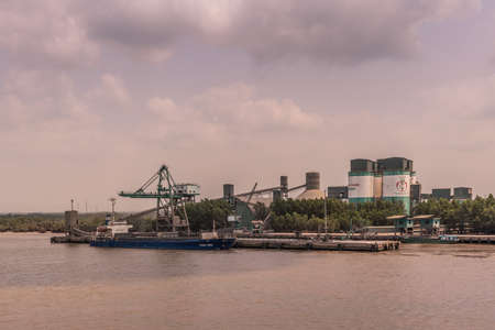 Long Tau River, Vietnam - March 12, 2019: Nhon Trach area. INSEE Cement station with silos, conveyor belt, and its quay where Tuan Minh boat is unloaded. under cloudscape. Brown river water.のeditorial素材