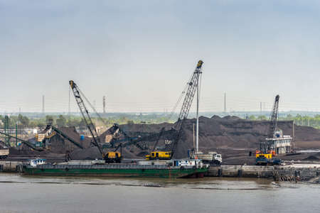 Long Tau River, Vietnam - March 12, 2019: Phuoc Khanh area. Coal port with quays and its hills of the black stuff. Tall crane and pumping trucks underlight blue sky. Long green boat is unloaded.のeditorial素材