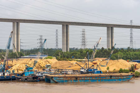 Long Tau River, Vietnam - March 12, 2019: Construction of Phuoc Khanh suspension bridge under silver sky, cut by many high voltage cables. Hills of yellow sand and blue pontoos on brown water. Green foliage.の写真素材