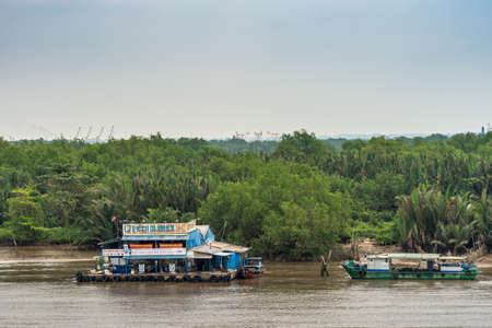 Ho Chi Minh City Vietnam - March 12, 2019: Petrolimex gas station for river boats with colorful banners on pontoon on Song Sai Gon river with green foliage screen in back.のeditorial素材