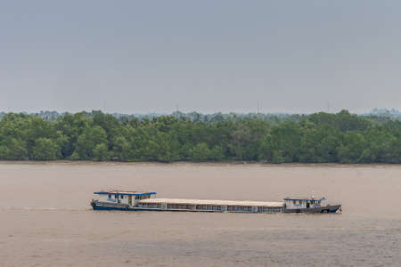 Long Tau River, Vietnam - March 12, 2019: Long hull-closed river barge sails on brown water and lies deep. Green belt separates from light blue sky.のeditorial素材