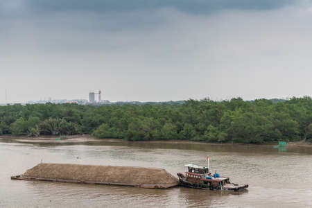 Ho Chi Minh City, Vietnam - March 12, 2019: Song Sai Gon river. Short tugboat pushes long barge filled with a long mount of sand. Brown water, green belt of jungle, and light blue sky.のeditorial素材