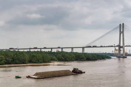 Ho Chi Minh City, Vietnam - March 12, 2019: Song Sai Gon river. Short tugboat pushes long barge with sand. Phu My bridge in back. Brown water, green belt of jungle, and light blue sky.のeditorial素材