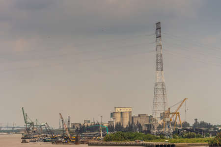 Long Tau River, Vietnam - March 12, 2019: Phuoc Khanh area. Chaotic photo of coal handling plant, with silos and cranes under tall high voltage pylon. Some green foliage in front under blue cloudscape.のeditorial素材