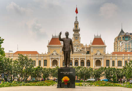Ho Chi Minh City, Vietnam - March 12, 2019: Downtown. Town Hall or Peopleâs Committee of HCMC French style building with clock tower and red roofs behind bonze statue of Ho Chi Minh himself under light blue sky, green foliage.のeditorial素材