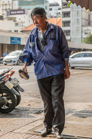 Ho Chi Minh City, Vietnam - March 12, 2019: Downtown street scenery. Closeup of older dreaming or thinking man dressed in black pants and blue company uniform vest strolls holding a drinking cup.のeditorial素材