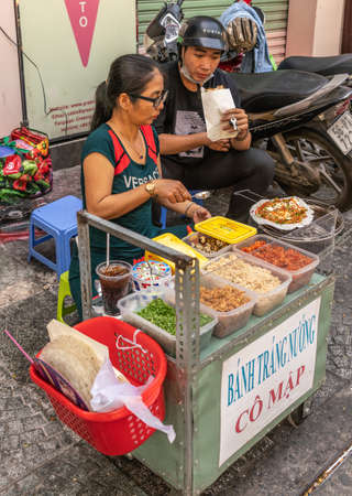 Ho Chi Minh City, Vietnam - March 12, 2019: Downtown street scenery. Ambulant female vendor sells fast food off push cart and grill. Client present and motorcycles.のeditorial素材