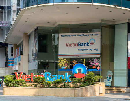 Ho Chi Minh City, Vietnam - March 12, 2019: Downtown street scenery. VietinBank office building entrance with big advertisement poster and red-=blue sign set up in green plant enclosure.のeditorial素材