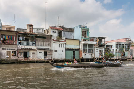Cai Be, Mekong Delta, Vietnam - March 13, 2019: Along Kinh 28 canal. 2 guys on low motorized boat on brown water passing in front of row of dwellings under blue cloudscape.のeditorial素材