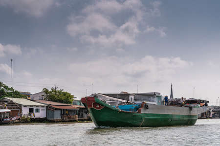 Cai Be, Mekong Delta, Vietnam - March 13, 2019: Along Kinh 28 canal. Green barge loaded with blue pipes sails under blue cloudscape. Poor housing on stilts on shoreline.のeditorial素材