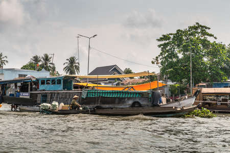 Cai Be, Mekong Delta, Vietnam - March 13, 2019: Retailer buyer on his sloop passes old large barge of supplier at floating wholesale market under gray cloudscape. Tarps and green foliage add color.のeditorial素材