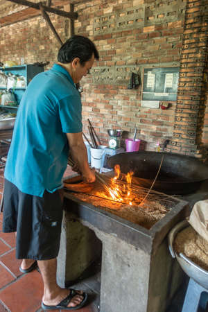 Cai Be, Mekong Deltal, Vietnam - March 13, 2019: man in blue shirt puts twigs on fire to light up his black vat to pop rice kernels against brown brick wall.のeditorial素材