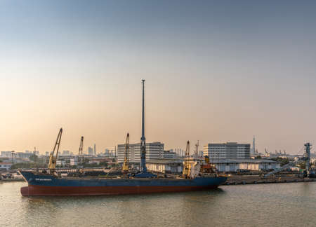 Ho Chi Minh City, Vietnam - March 13, 2019: Song Sai Gon river at sunset. Blue-red Ocean Bright sea cargo vessel docked and worked on by tall blue crane. Coils of black steel wire on yard. Buildings on far horizon.のeditorial素材
