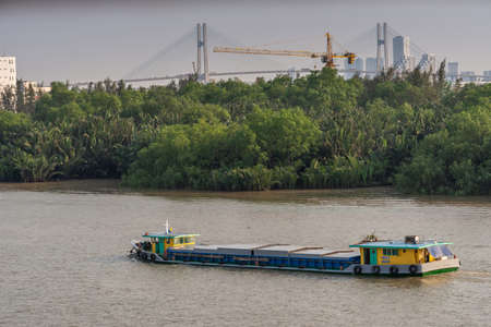 Ho Chi Minh City, Vietnam - March 13, 2019: Sunset sky shot over Song Sai Gon river. Front steering barge with green housing on stern has closed hold. Green belt and Phu My bridge on horizon.のeditorial素材