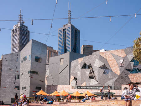 Melbourne, Australia - November 17, 2009: Federation square baking in sun under blue sky in front of ACMI modern architecture building. People and orange umbrellas. Skyscrapers and cables.のeditorial素材