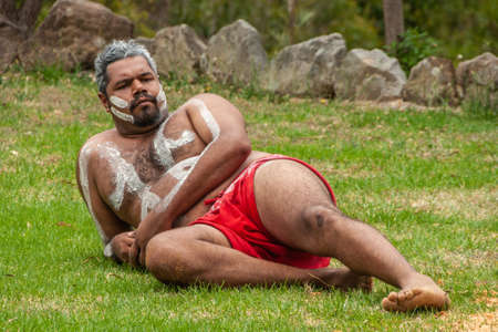 Newcastle, Australia - December 10, 2009: Closeup of Aboriginal man relaxing on green lawn. White stripes on half-naked body and red groin cloth.のeditorial素材