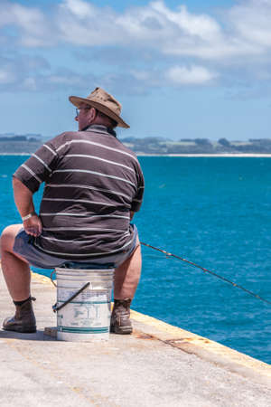 Stanley, Tasmania, Australia - December 15, 2009: Corpulent man dressed in shorts and brown T-shirt sits on white paint bucket while fishing with rod in blue sea under blue cloudscape.のeditorial素材