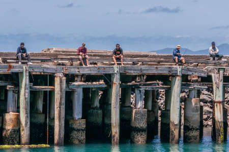 Stanley, Tasmania, Australia - December 15, 2009: Row of 5 men sitting on gray-brown wooden pier fishing in azure sea with rods under blue cloudscape.のeditorial素材