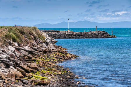 Stanley, Tasmania, Australia - December 15, 2009: Rocky coastline and breakwater of the port in blue seawater under blue cloudscape with dark hills on other side of bay.の写真素材