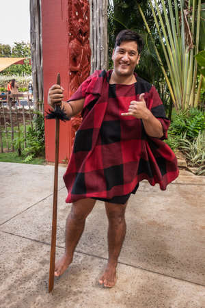 Laie, Oahu, Hawaii, USA. - January 09, 2020: Polynesian Cultural Center. Closeup of young male Maori warrior showing the Shaka sign. Black and red garb. Back is green vegetation and maroon totem sculptures.のeditorial素材