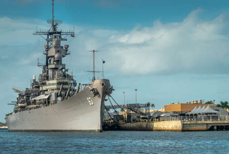 Oahu, Hawaii, USA. - January 10, 2020: Pearl Harbor. Bow of Gray USS Missouri docked and used as historic museum on blue water and under blue cloudscape. Brown buildings on shore.のeditorial素材
