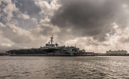 Oahu, Hawaii, USA. - January 10, 2020: Pearl Harbor. Long shot of Gray Abraham Lincoln aircraft carrier docked under full rain dark cloudscape on dark gray water.のeditorial素材