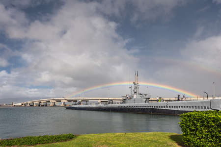 Oahu, Hawaii, USA. - January 10, 2020: Pearl Harbor. Rainbow straddles long submarine USS Bowfin sticking out of gray water under blue cloudscape. Ford Island bridge lines up with boat. Green foliage up front.のeditorial素材