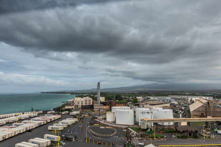 Kahului, Maui,, Hawaii, USA. - January 13, 2020: Huge dark cloud in full gray cloudscape hangs over the port area with its fuel tanks and shipping containers. Azure ocean and green zone in back.のeditorial素材
