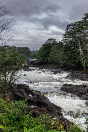 Hilo, Hawaii, USA. - January 14, 2020: Wailuku River surrounded by green trees and plants above Rainbow Falls runs over black volcanic rocks under rainy cloudscape.の写真素材